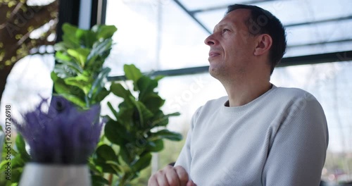 A man sits calmly at a sunlit table in a cafe, absorbed in reading as vibrant plants surround him. The relaxing ambiance highlights his focus and the day's tranquility