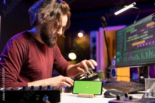 Foto Composer singer mixing tunes on soundboard in home studio, editing next to a smartphone with greenscreen layout