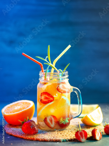 Infused detox water with orange, strawberry, lemon and rosemary. A cold homemade summer drink, sliced pieces of citrus fruits and berries. Blue background