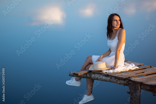 A beautiful young woman in a summer white dress sits by the water on a wooden bridge. The blue sky is reflected in the water , copy space . Summer vacation