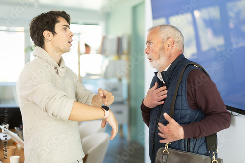 Картината върху платно Young man sternly scolds elderly male colleague for being late for work in offic
