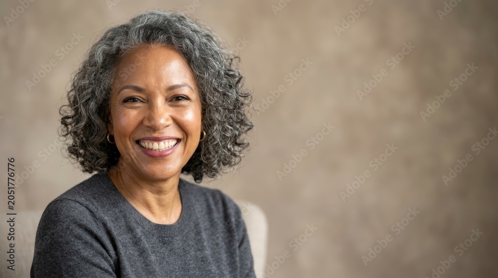 Fototapeta premium Portrait of a happy middle aged woman with curly grey hair smiling. Cheerful mature lady looking at camera. Concept of wellness, beauty, confidence, aging gracefully and positive mindset.
