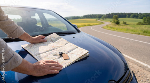 Man holding paper map with car key on hood of blue vehicle. Rent a car concept, traveler planning route on road trip. Summer vacation, transport navigation and travel agency service.