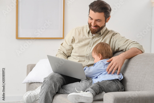 Working father with laptop and his little baby on sofa during parental leave ...
