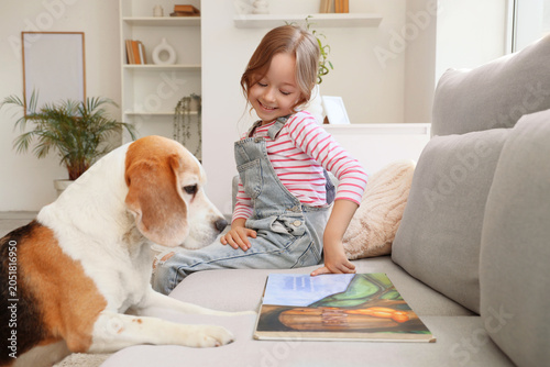 Foto Cute little girl with Beagle dog reading book on sofa in living room