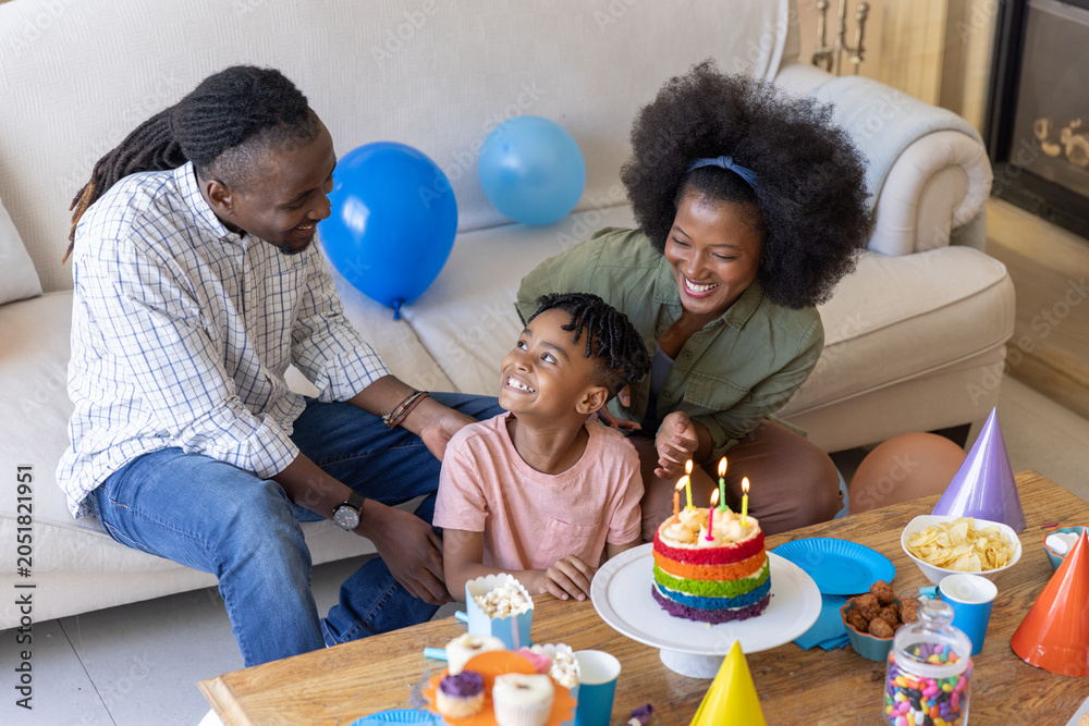 Obraz premium Diverse family celebrating birthday in living room at coffee table with rainbow cake and balloons