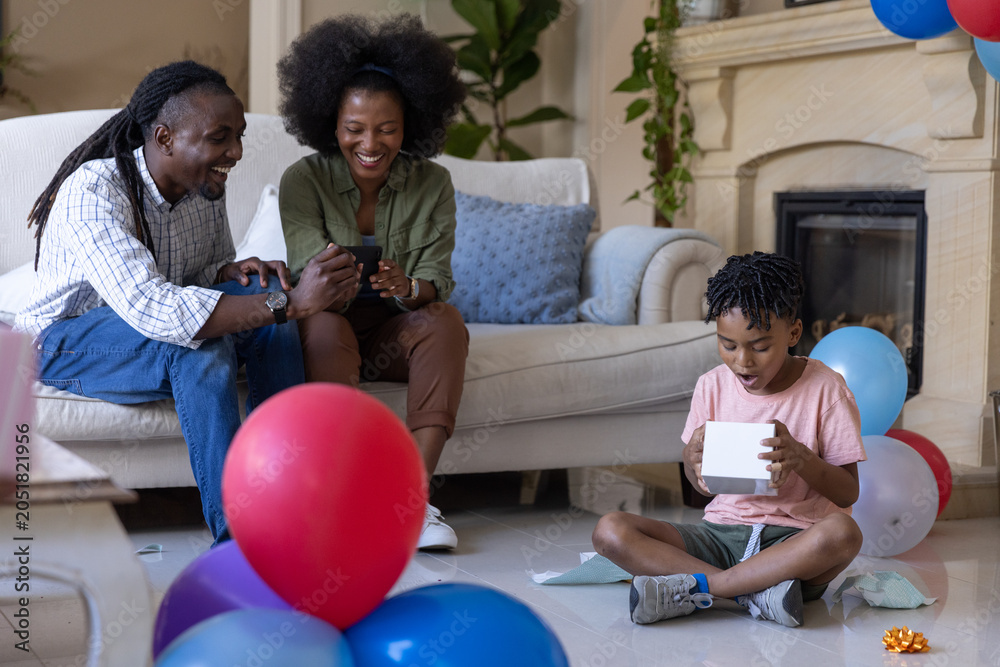 Obraz premium African American family sitting in living room smiling at phone, child opening gift box, copy space