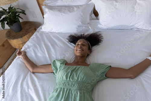 Female lying across queen bed in bedroom beside headboard with potted plant, smiling eyes closed