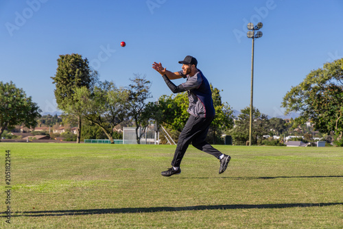 Red cricket ball hovering above green sports field during sunny practice near floodlight mast