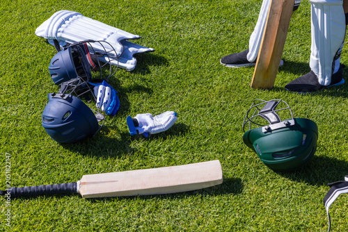 Cricket bat, helmets, gloves and pads are lying scattered on well-trimmed grass under bright sun