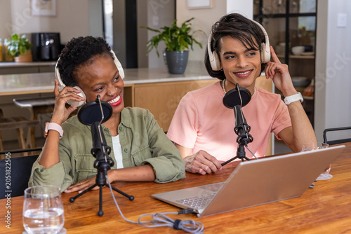 Diverse co-hosts sitting in home kitchen recording with laptop, microphones and headphones, smiling