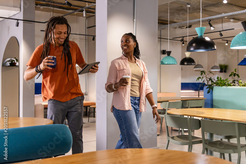 African American coworkers walking, chatting in lounge with casual wear, tablet, mug and paper cup
