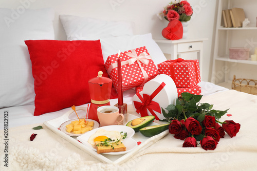 Tasty breakfast with roses and gifts on bed in room