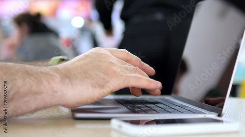 Hand types on laptop keyboard. Fingers press keys smoothly. Background shows blurred people in cafe. Laptop sits on wooden surface. White phone rests nearby. Shot focuses on typing action