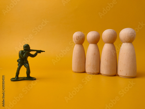 A green plastic toy soldier aims a rifle at a row of four wooden peg figures against a yellow background