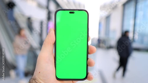Hand holds smartphone with green screen. Background shows blurred public space. Person appears calm, focused. Ideal for app demos, digital ads, or UI mockups