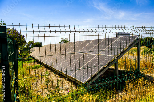 Photovoltaic solar panels in a countryside near highway surrounded by fence under sunny blue sky, renewable energy carbon footprint reduction