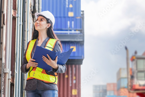 Female logistics supervisor holding a clipboard inspecting cargo containers at a busy port, Professional woman in safety gear and hard hat monitoring shipping yard operations outdoors