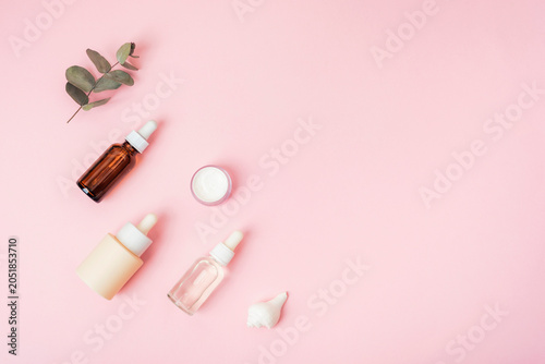 Skincare products with serum bottles, cream jar and eucalyptus branch arranged on pink background with copy space. Top view