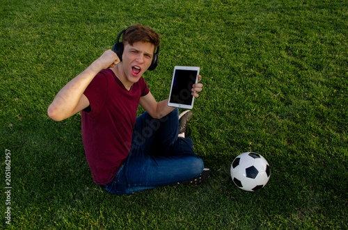 Blonde teenager sitting on the fresh cut grass holding tablet with empty screen , looking at camera and cheering 