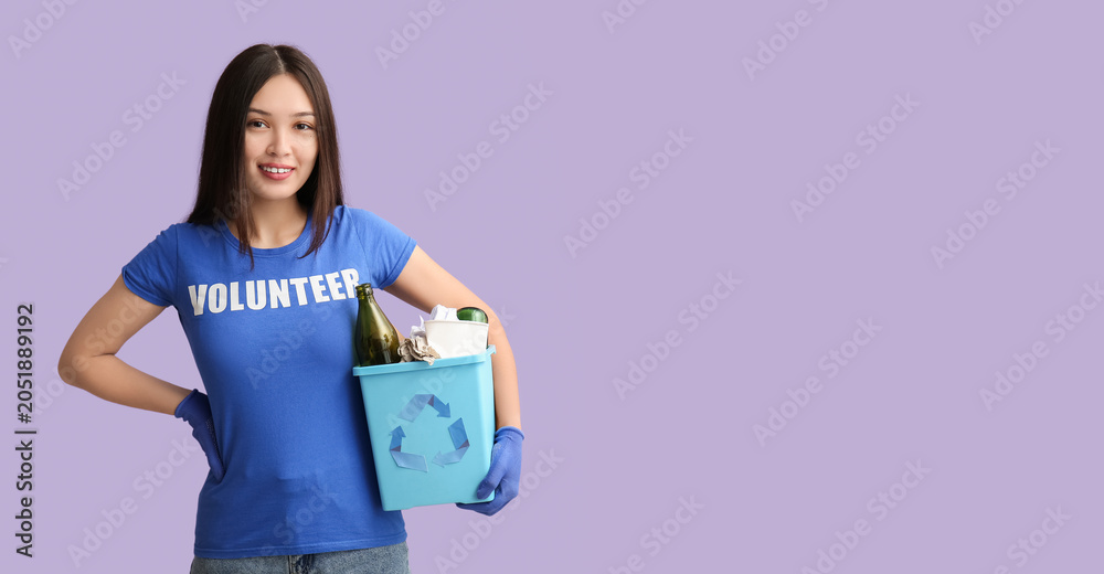 Fototapeta premium Young female volunteer holding recycle bin with garbage on lilac background