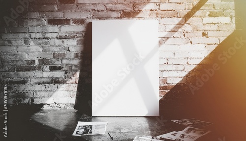 Blank white canvas leaning against a rustic brick wall with strong shadows and scattered papers on the floor.
