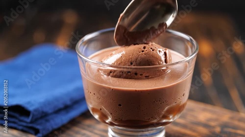 Spoonful of airy chocolate mousse being lifted from a glass bowl on a rustic wooden table with a blue napkin