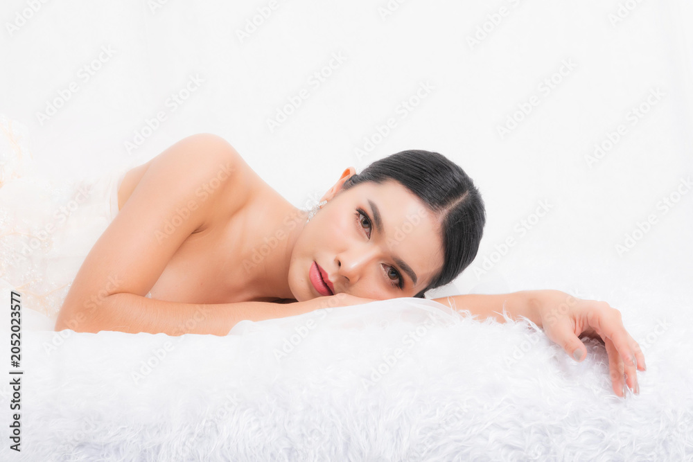 Fototapeta premium Portrait capturing an intimate moment with a beautiful Asian bride in a white gown, lying down gracefully with her hands clasped studio shot on white background.