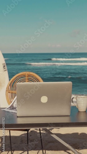 A modern laptop and a cup of coffee sit on a table at a sunny tropical beach. The beautiful blue ocean waves in the background create a perfect remote workspace for a digital nomad.