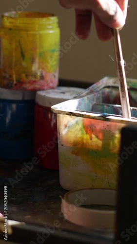 Close-up of single flat paint brush with orange acrylic paint on bristle tip balanced on edge of messy metal palette in dark studio
