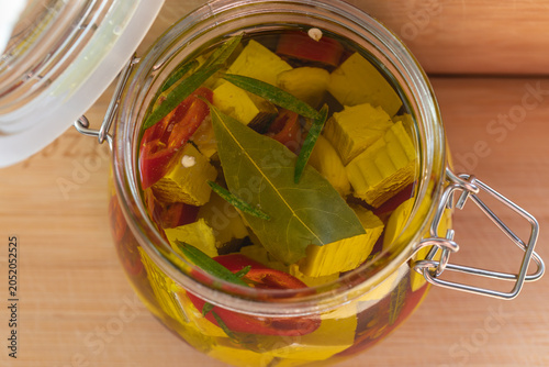 Close up top view of an open wire-bail glass jar filled with marinated yellow feta or tofu cubes, bay leaves, chili, and rosemary in olive oil.