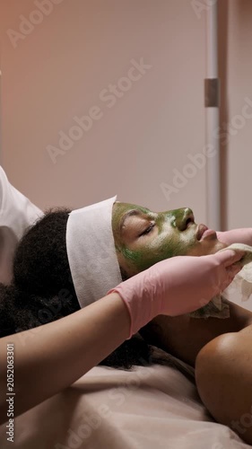 Close up of a professional cosmetologist removing green detox or seaweed mask from african american client face using a wet wipe in a salon. Skincare routine. Vertical