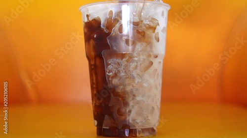 4K Pouring rich espresso into plastic cup of milk and ice blending into delicious iced latte, Refreshing cold caffeine drink isolated on vibrant orange background.