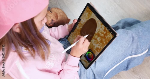 Child wearing pink cap drawing cute cartoon capybara on digital tablet screen with stylus. Creative educational graphic design software activity for early child development at home. Real indoor summer