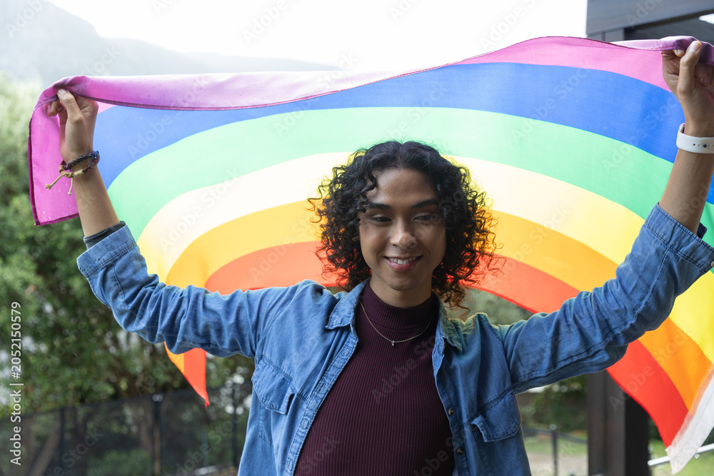 Obraz premium Non-binary adult standing on balcony holding rainbow flag behind shoulders, wearing denim jacket