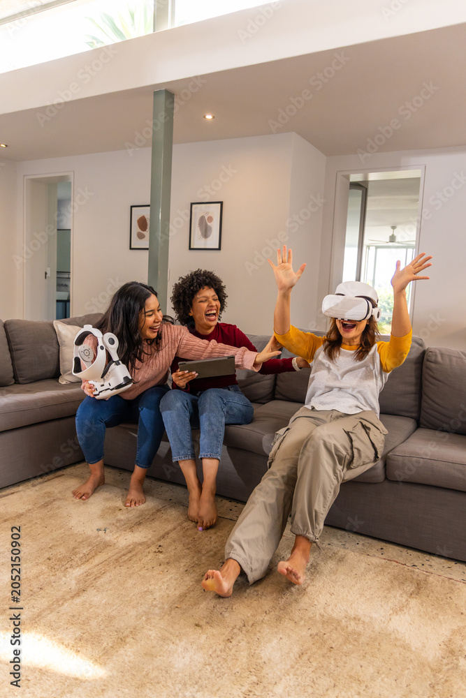 Naklejka premium Diverse female friends laughing on sectional sofa in living room with VR headset, tablet, toy robot