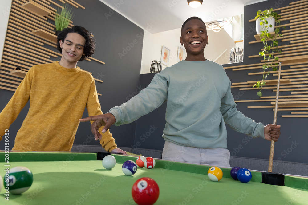 Naklejka premium Diverse teenage male friends playing on green pool table in modern rec room with cue stick