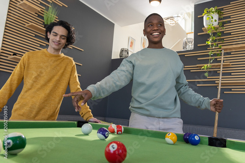 Diverse teenage male friends playing on green pool table in modern rec room with cue stick