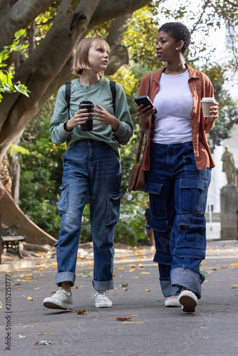 Diverse female friends walking on park path holding travel mug, coffee cup, smartphone, backpacks