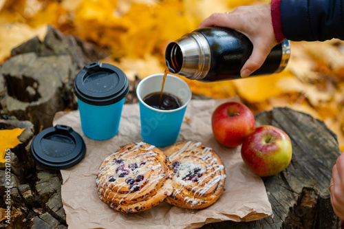 Autumn picnic concept - female hand pouring hot coffee into cup on tree stump with pastries and apples among colorful leaves