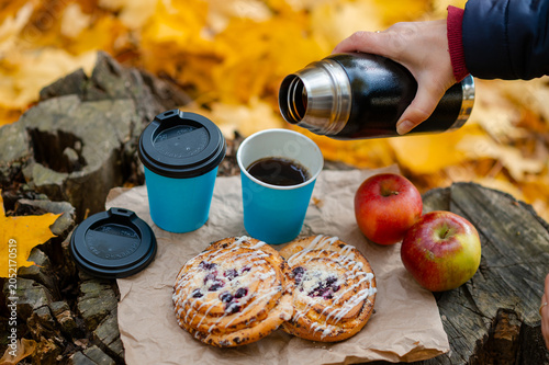 Autumn picnic concept - female hand pouring hot coffee into cup on tree stump with pastries and apples among colorful leaves