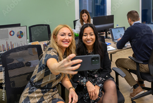 Diverse business women friends smiling and taking a selfie with phone together in a coworking office while colleagues work on computers in the background, showing collaboration and friendship