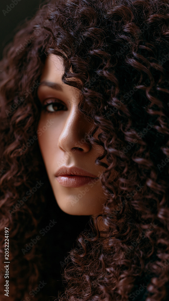 Fototapeta premium Thoughtful woman with curly hair looking through a window in soft natural light with a contemplative expression.