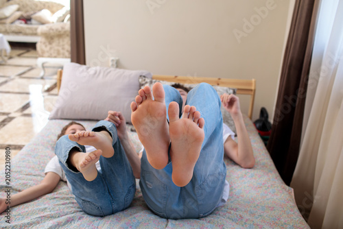Two children lying on a bed with bare feet raised toward camera, both in jeans, playful indoor home scene