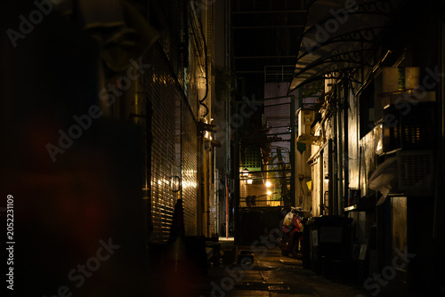 Photos A dark and narrow back alley at night in Wan Chai, Hong Kong, with warm glowing lights receding into the distance, a lone figure crouching beside a parked scooter