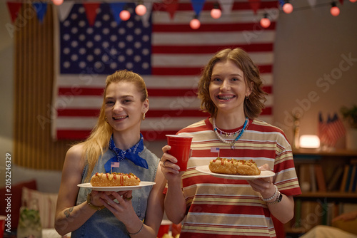 Canvas Print Portrait of Caucasian teenage girls holding hot dogs and drink, celebrating Independence Day at home with USA flag backdrop