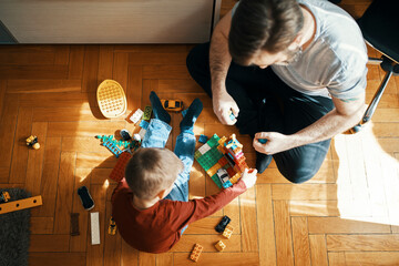 Father and son sitting on the floor playing together with building bricks, top view