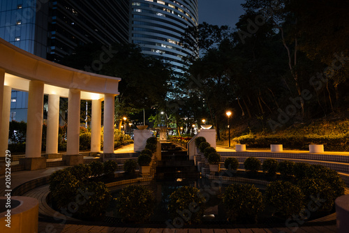 Obraz na plátně Night view of the illuminated Olympic Square amphitheatre in Hong Kong Park, Central, with its classical columns, ornamental pool and topiary garden framed by towering skyscrapers and lush trees