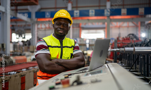Konstfotografi Industrial Operator at Work: A focused industrial operator, wearing a yellow hard hat and safety vest, stands confidently in a factory setting, his arms crossed, with a laptop open before him