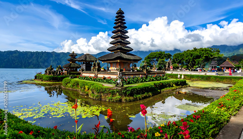 Iconic Balinese water temple complex on serene lake reflecting blue sky and white clouds, lush greenery and flowers in foreground